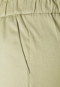 Detailní záběr na světle zelenou látku se shromážděným elastickým pásem v pase a vertikální boční kapsou na kalhotách nebo sukni.