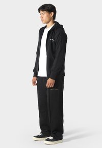 Felpa nera con zip e tasche frontali, decorata con lettering bianco, abbinata a pantaloni della tuta neri con ricami dorati. Il modello indossa una maglietta bianca.