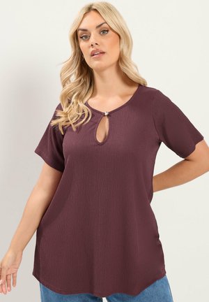 Woman with blonde hair wearing a short-sleeve textured purple top with a keyhole neckline and blue jeans against a plain light background.
