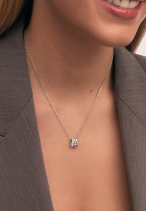 Silberne Halskette mit einer Kette, die einen Anhänger aus drei verwobenen Ringen zeigt, von denen einer mit kleinen, klaren Steinen verziert ist und auf einer strukturierten Oberfläche ruht.