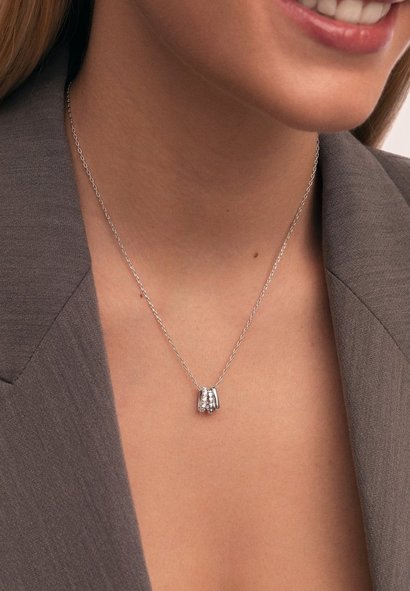 Silberne Halskette mit einer Kette, die einen Anhänger aus drei verwobenen Ringen zeigt, von denen einer mit kleinen, klaren Steinen verziert ist und auf einer strukturierten Oberfläche ruht.