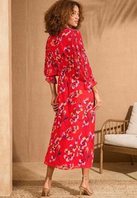 Rochie maxi roșie cu flori, cu mâneci lungi pliate și talie strânsă. Prezintă un material moale, un model floral multicolor și un design fluid.