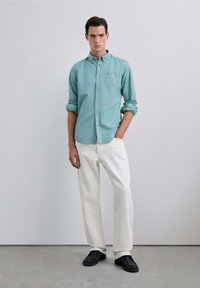 Camisa de botones en un azul claro con un bolsillo en el pecho, mangas enrolladas, combinada con pantalones rectos blancos y zapatos negros tipo slip-on.