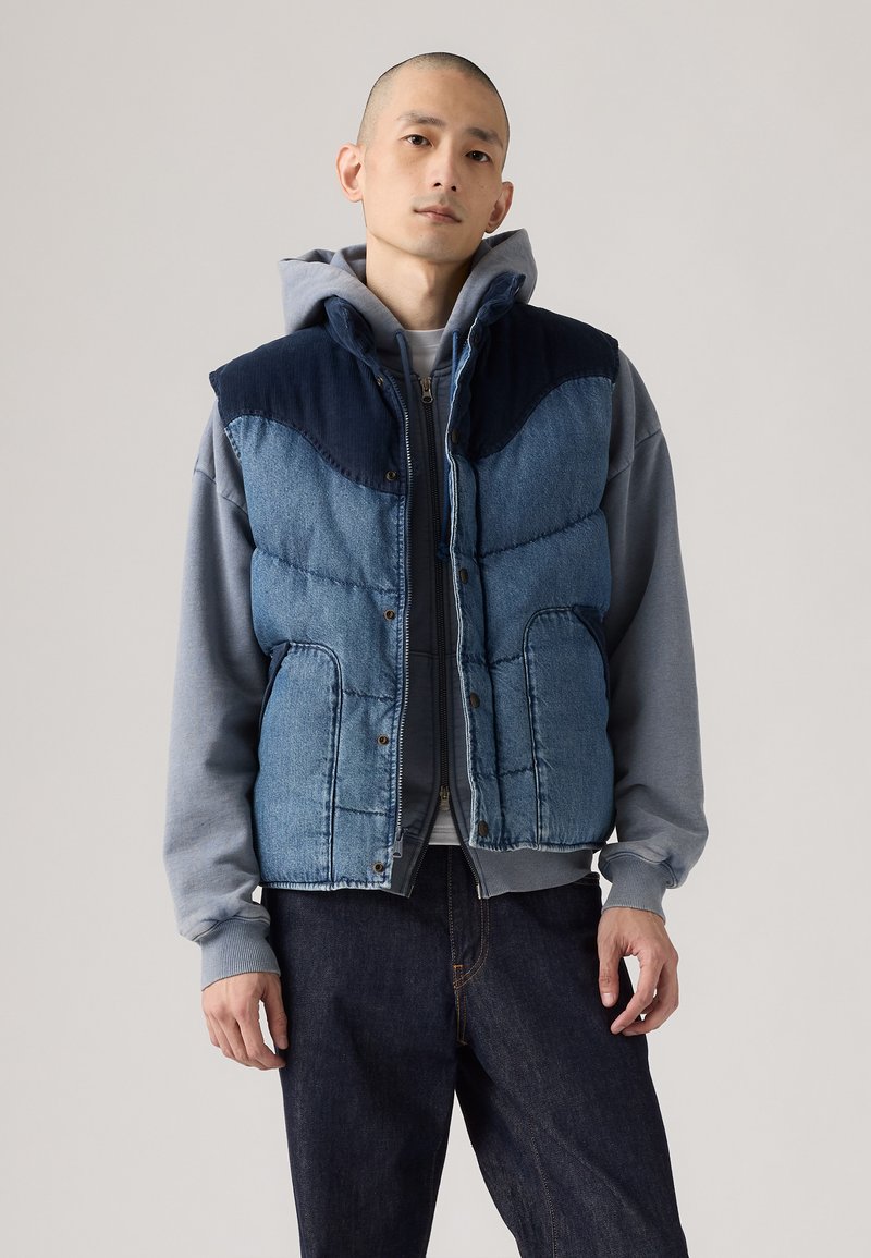Denim puffer vest med en mørkeblå krave, der har quiltet tekstur, trykknapper og forlommer, lagdelt over en grå hoodie.