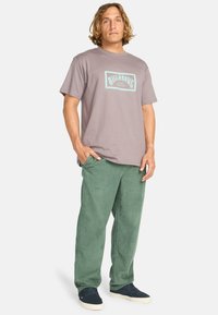 Gray t-shirt with turquoise graphic, paired with green corduroy pants. Model stands casually with hands in pockets, wearing navy sneakers.
