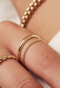 Three gold rings on a hand: a smooth band, a beaded band, and a twisted band, all set against a soft, muted background.