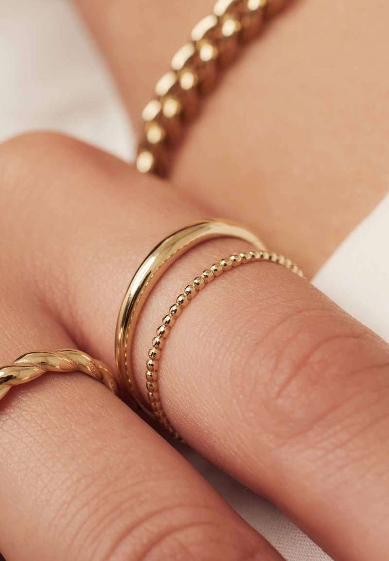 Three gold rings on a hand: a smooth band, a beaded band, and a twisted band, all set against a soft, muted background.