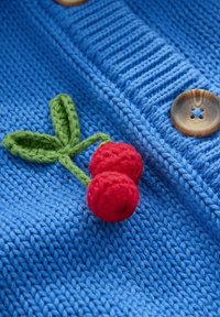 Cardigan en tricot bleu avec deux cerises en crochet rouges et des feuilles vertes, et des boutons marron clair. Tissu texturé avec des détails côtelés.