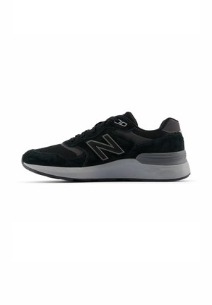 Zapatilla New Balance negra y gris con parte superior de malla y ante, cierre de cordones y suela acolchada, mostrada de lado sobre fondo blanco.