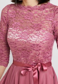 Krajkový top v mauve s vykrojeným výstřihem a tříčtvrtečními rukávy, doplněný jednobarevnou mauve sukní a saténovým páskem.