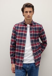 Rot-kariertes Hemd in Marineblau mit Stehkragen zum Knöpfen, langen Ärmeln und weißen Knöpfen; getragen über einem weißen T-Shirt und blauen Jeans.