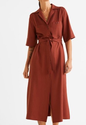 Robe couleur rouille avec col chemise, manches courtes, deux poches poitrine, fermeture à boutons et ceinture à la taille, présentant une coupe fluide mi-longue.