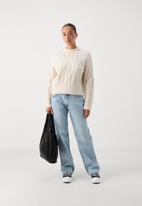 Πουλόβερ χοντρής πλέξης σε κρέμα απόχρωση με διαμαντένια μοτίβα, συνδυασμένο με ανοιχτό μπλε παντελόνι με φαρδιά πόδια και μαύρη πλισέ τσάντα χειρός; λευκά αθλητικά παπούτσια με μαύρες ρίγες.