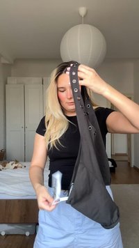 Schwarzfarbenes Top mit einem Schulterdesign, verziert mit Ösen und aus weichem Stoff, gehalten von einer Person in einem Raum mit hellen Möbeln und Bettwäsche.