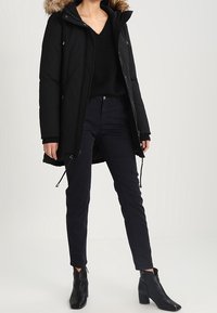 Zwarte pufferjas met een gevoerd capuchon van nepbont, ritssluiting, getailleerde taille, gecombineerd met donkere jeans en enkellaarsjes. Gladde textuur, middelgrote lengte.
