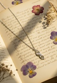 Silverkedja med ett pendang som har en oändlighetsform, med en slät yta och en liten gnistrande detalj, placerad på en handskriven bakgrund.