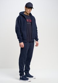Sudadera azul marino con cremallera y bolsillos frontales, combinada con una camiseta azul marino a juego y pantalones de chándal. Presenta tejido texturizado y puños elásticos.