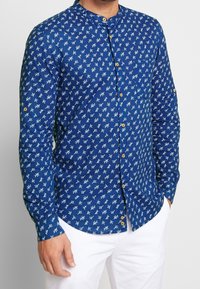 Blå skjorta med mandarinkrage, med ett litet vitt blommönster och rundad nederkant. Tillverkad av lättviktsmaterial.