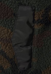 Närbild av ett mörkt kamouflagemönstrat fleece-material med en sydd rektangulär svart nylonlapp.