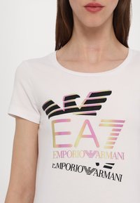 Bílý bavlněný tričko s barevnými logy EA7 a Emporio Armani. Design zahrnuje vrstvené pruhy v růžové, černé a v odstínech gradientu.