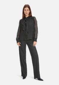 Schwarze Bluse mit Polka-Dots aus transparenter Stoff, mit Rüschenkanten und einem Schleifen-Ausschnitt, kombiniert mit schwarzen weit geschnittenen Hosen und spitzen Schuhen.