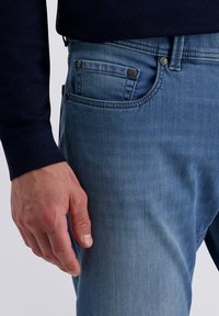 Mann in blauen Jeans und einem dunkelblauen Pullover, mit der linken Hand entspannt an seiner Seite.