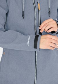 Gray outdoor jacket with a textured finish, front zipper, adjustable cuffs featuring Velcro, and a logo on the sleeve.