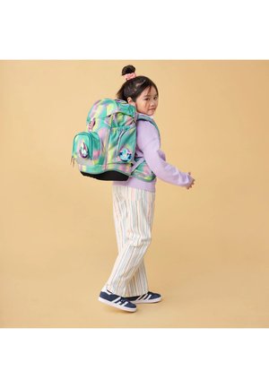 Young girl wearing pastel sweatshirt and striped pants carries large colorful backpack, looking back over shoulder against beige background.