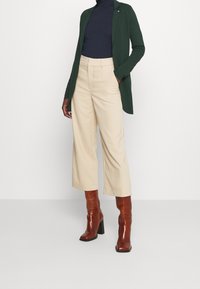 Henkilö, joka on pukeutunut beigeksi värjättyihin leveisiin lyhyisiin housuihin, tumman vihreään bleiseriin, laivastonsiniseen poolopaitaan ja ruskeisiin korkeakorkoisiin nahkasaappaisiin, seisoo valkoista taustaa vasten.