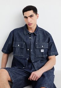 Short-sleeve denim shirt in dark blue with two chest pockets, button front, and Lee logo patch. Coordinating denim shorts are also visible.
