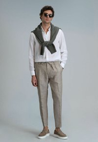 Chemise en lin blanche, pantalon rayé beige clair, pull en tricot vert olive drapé sur les épaules, chaussures à enfiler marron avec semelles blanches.