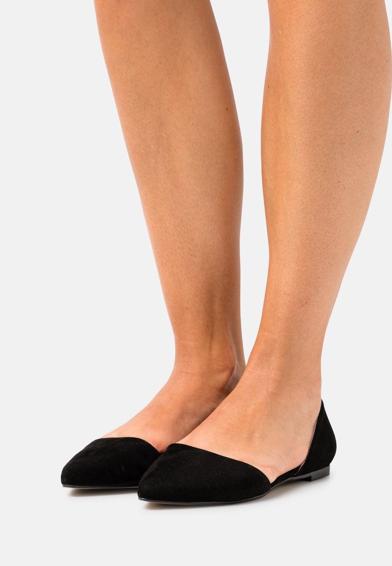 Jambes portant des ballerines noires à bout pointu et à petits talons, debout sur un fond blanc.