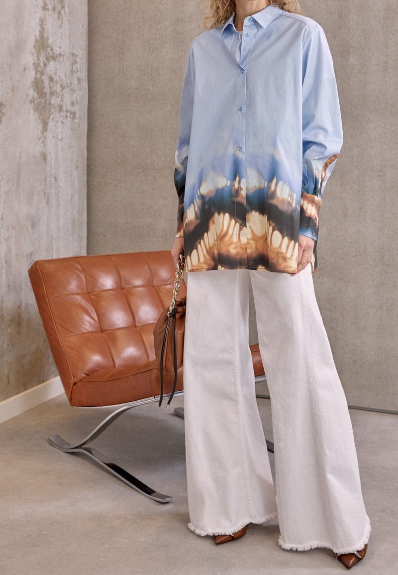 Light blue button-up shirt with patterned gradient in brown and blue, paired with white wide-leg trousers and brown heeled shoes.