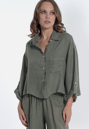 Donna che indossa una camicia verde oliva morbida con bottoni e pantaloni abbinati, che guarda leggermente di lato su uno sfondo semplice.
