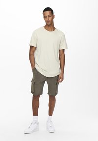 Helles beigefarbenes T-Shirt, kombiniert mit olivgrünen Cargo-Shorts mit seitlichen Taschen und ausgefranstem Saum, vervollständigt mit weißen Sneakern und Knöchelsocken.
