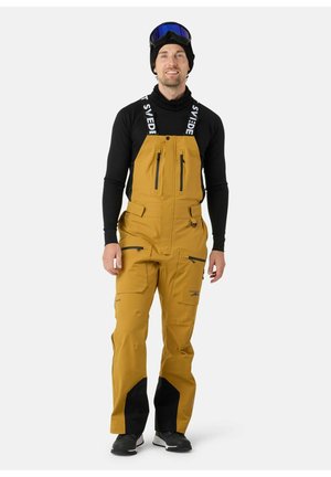 Geel skibroek met zwarte accenten en ritsen. Beschikt over verstelbare banden en een gladde textuur. Draagt over een zwart shirt met lange mouwen.