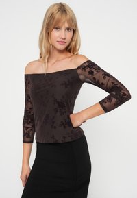 Off-shoulder topp i mørk brun med gjennomsiktige lange ermer som har et blomstermønster. Stoffet har en glatt tekstur. Kombinert med et svart skjørt.