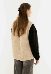 Beige Fleece-Weste mit schwarzen langen Ärmeln; strukturierter, oversize Schnitt. Langes brünettes Haar, teilweise mit einer schwarzen Klammer zurückgehalten. Graue Hose.