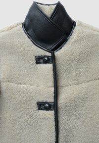 Manteau en peau lainée avec un corps texturé crème, un col en cuir noir, et une fermeture à boutons avec un accent contrastant noir.