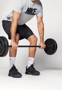 T-shirt gris Nike avec logo audacieux, shorts noirs, chaussures de sport noires et chaussettes blanches à la cheville, tenant une barre noire avec des poids.
