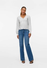 Cardigan tricoté gris clair avec un col festonné, une fermeture à boutons et des manches longues, associé à un jean bleu évasé et des sandales noires.