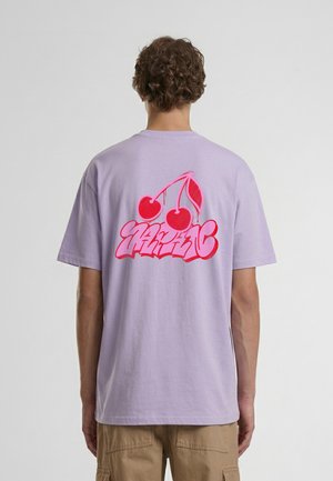 Personne portant un t-shirt violet clair avec des cerises rouges éclatantes et un texte de style graffiti dans le dos, associé à un short cargo beige.