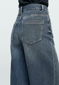 Vista posteriore di una persona che indossa jeans grigi in denim a vita alta con gamba dritta, cuciture visibili e tasche posteriori, su uno sfondo uniforme.