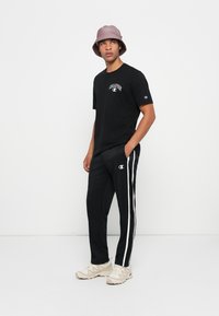 Mann in schwarzem Champion-T-Shirt und schwarzen Trainingshosen mit weißen Streifen, beigen Turnschuhen und einem rosa Buckethat, stehend vor einem weißen Hintergrund.