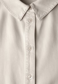 Camicia col beige chiaro con colletto e una texture liscia. Presenta una chiusura con bottoni a pressione con quattro bottoni abbinati e dettagli cuciti sul colletto.