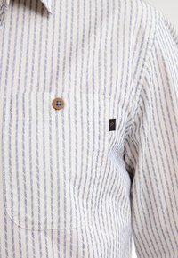 Camicia a righe bianca e azzurro chiaro, con taschino sul petto, dettagli con bottoni e piccola patch del logo vicino all'orlo. Tessuto leggero.