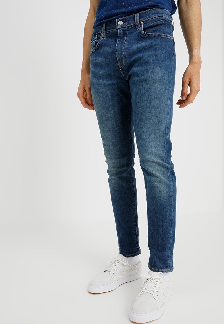 Homme portant un jean bleu slim et des baskets blanches, debout devant un fond uni.