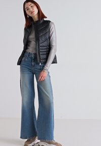Black padded vest over a gray long-sleeve top, paired with wide-leg faded blue jeans and beige sneakers. Smooth textures and layered design.
