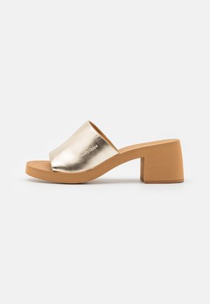 Mule sandale à bout ouvert avec empeigne dorée métallique et talon bloc épais couleur beige clair sur fond clair.