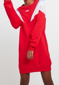 Robe sweat-shirt rouge en tissu doux, avec des rayures diagonales blanches et bleues sur les épaules et un col rond.